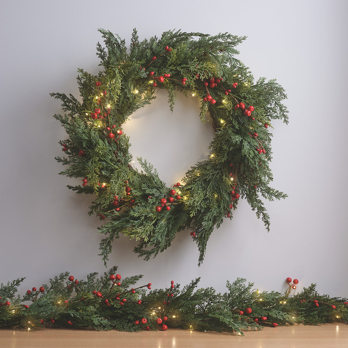 Weihnachtskranz mit roten Beeren und LED-Lichtern an weißer Wand mit Tannenzweigen