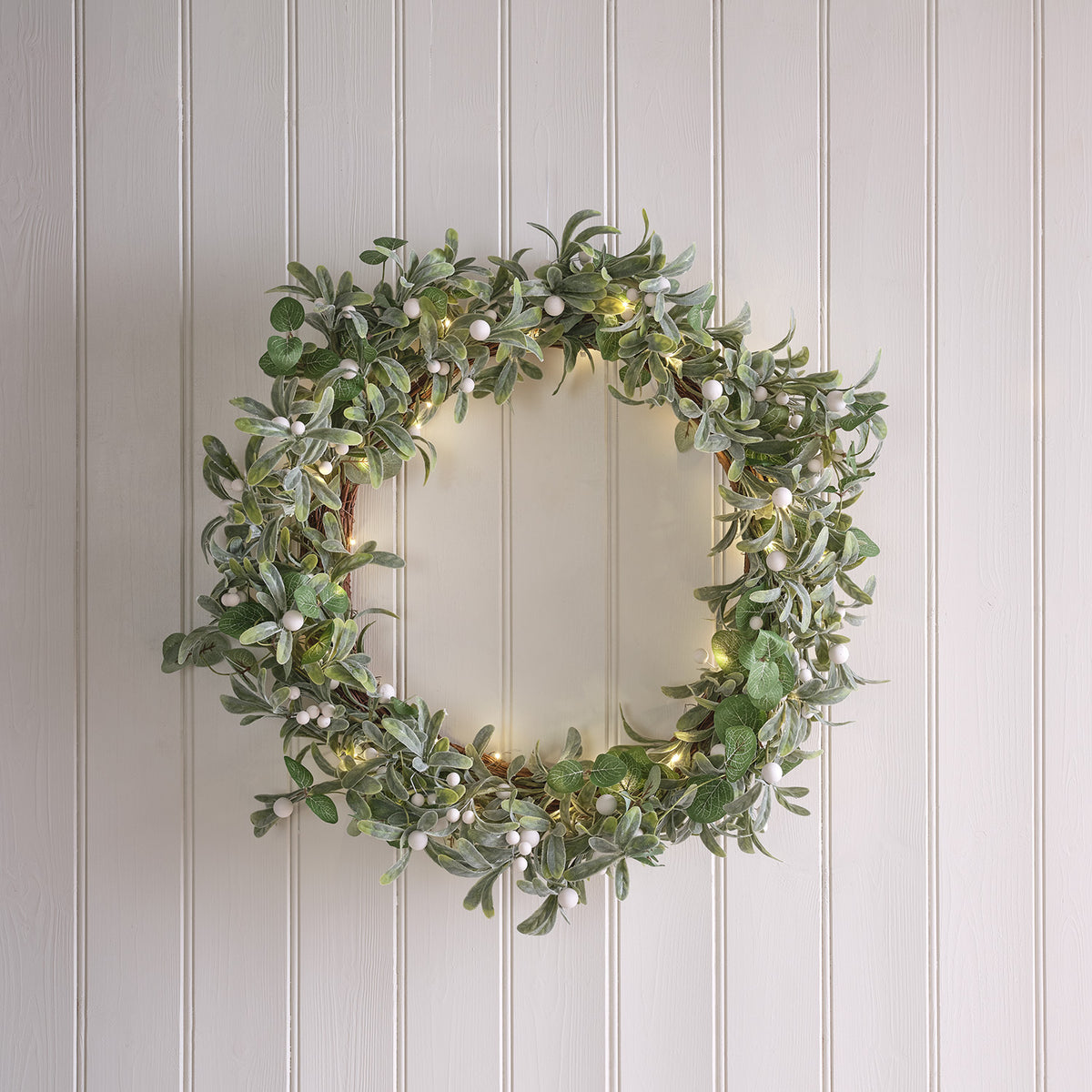 Beleuchteter Weihnachtskranz mit grünen Blättern und weißen Beeren an weißer Holzverkleidung