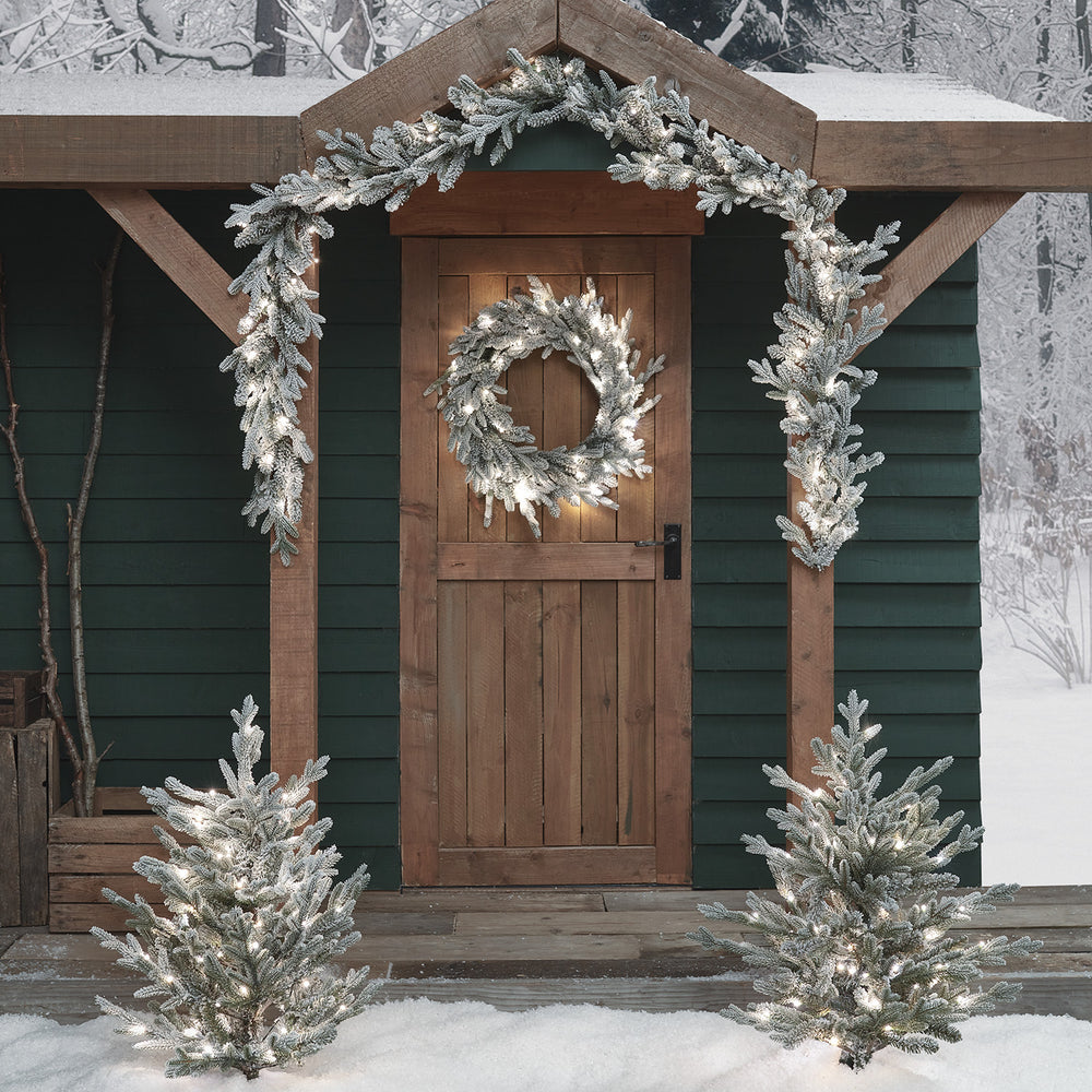 Verschneiter Weihnachtskranz und Girlande mit weißen LEDs an Holztür und Eingang