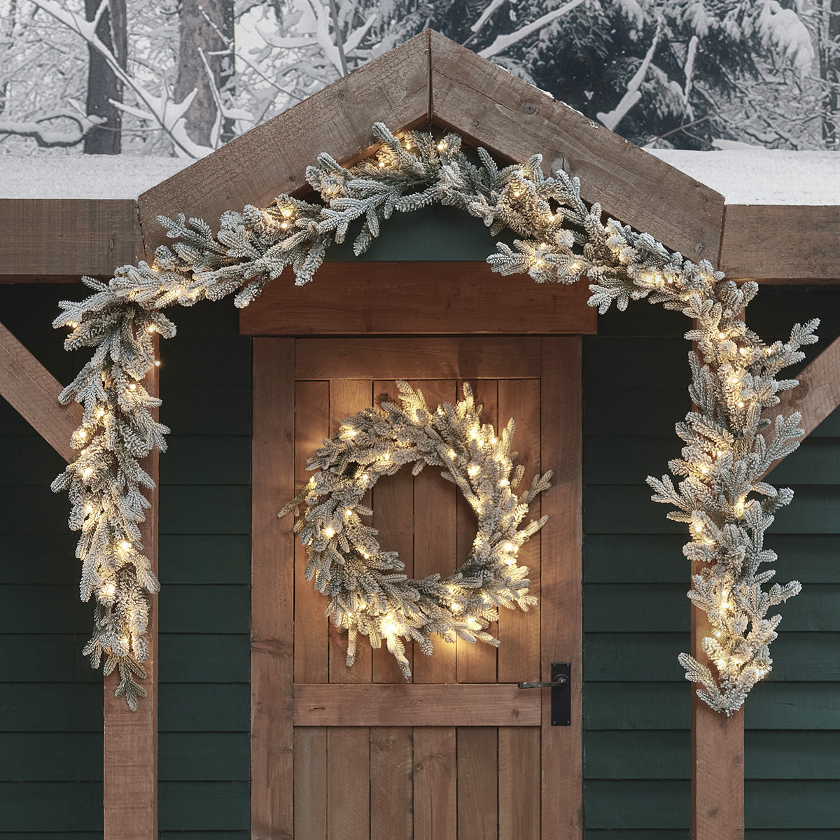 Verschneite Weihnachtsgirlande und Kranz mit warmweißen LEDs an Holztür und Dachgiebel