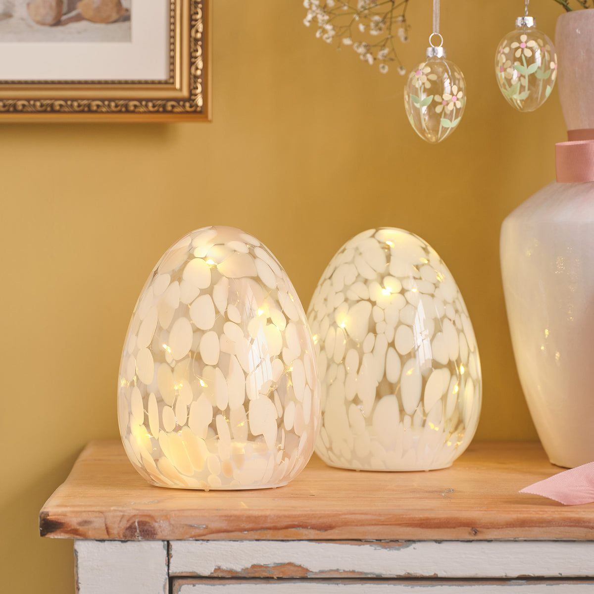 Zwei handgeblasene Glas-Ostereier mit warmweißen Mikro-LEDs und weißem Konfetti-Muster auf Holzbrett