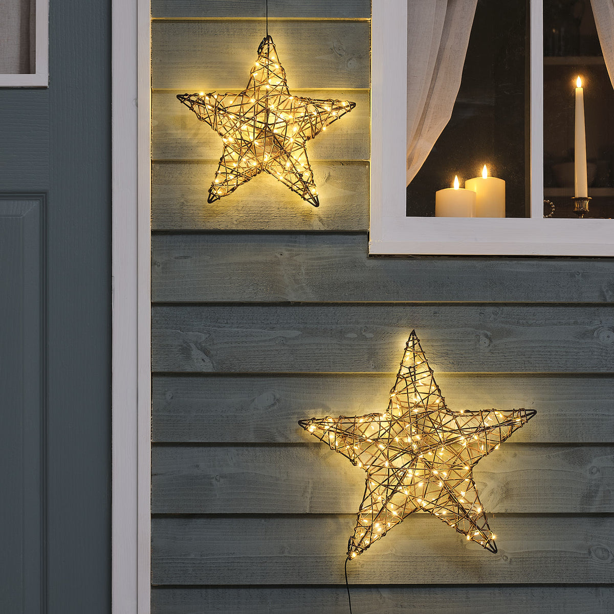 Zwei beleuchtete Rattan-LED-Sterne in 30 und 40 cm an grauer Holzfassade bei Dämmerung