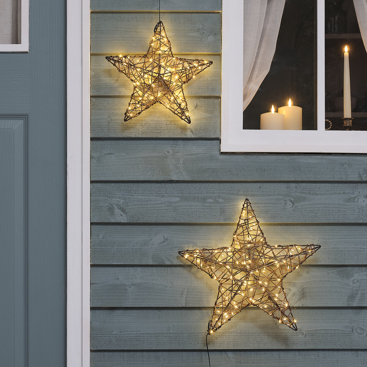 Zwei beleuchtete Rattan-LED-Sterne in 30 und 40 cm an grauer Holzfassade bei Dämmerung