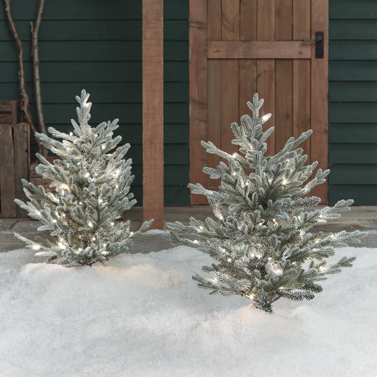 Zwei verschneite LED-Weihnachtsbäume mit weißen Lichtern im Schnee vor Holztür