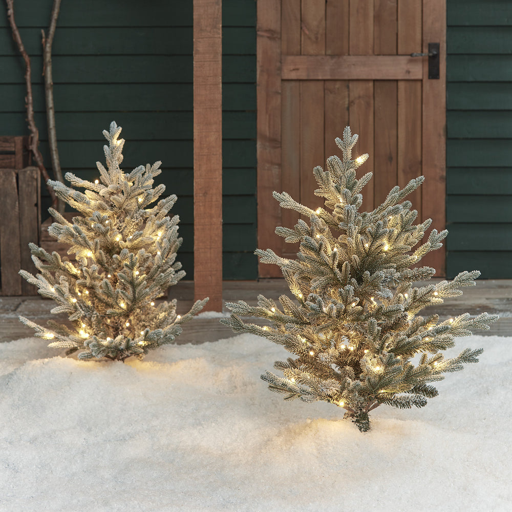 Zwei verschneite LED-Weihnachtsbäume mit warmweißen Lichtern im Schnee vor Holztür