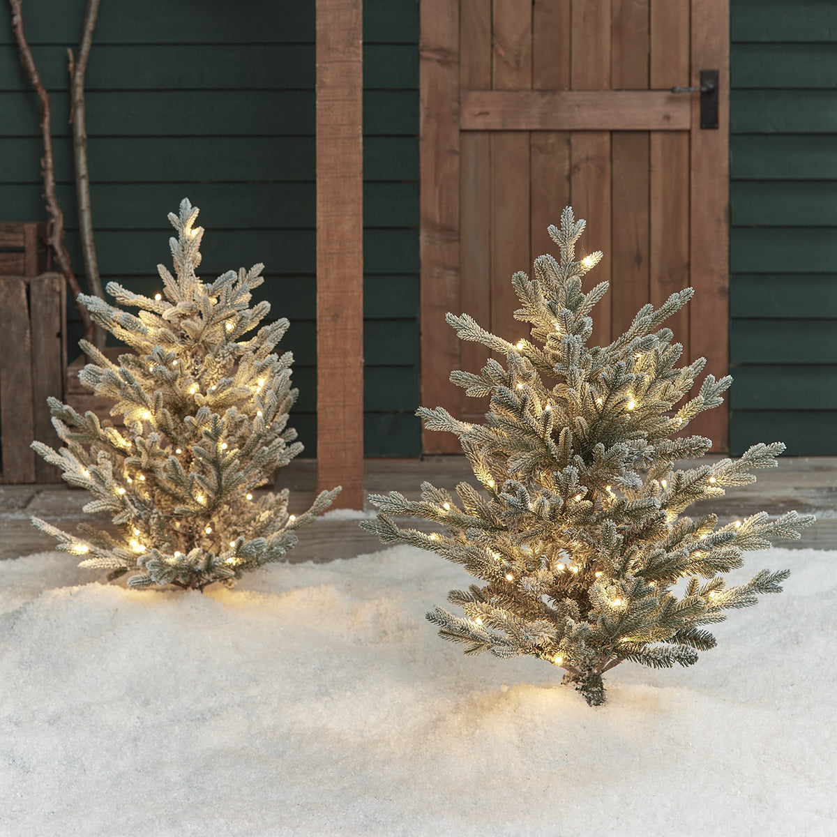 Zwei verschneite LED-Weihnachtsbäume mit warmweißen Lichtern im Schnee vor Holztür