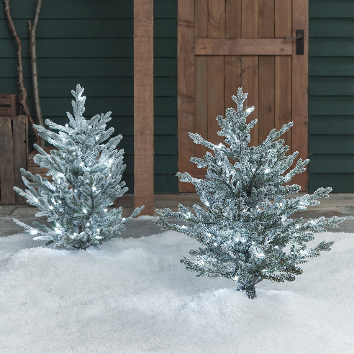 Zwei verschneite LED-Weihnachtsbäume mit kaltweißen Lichtern im Schnee vor Holztür