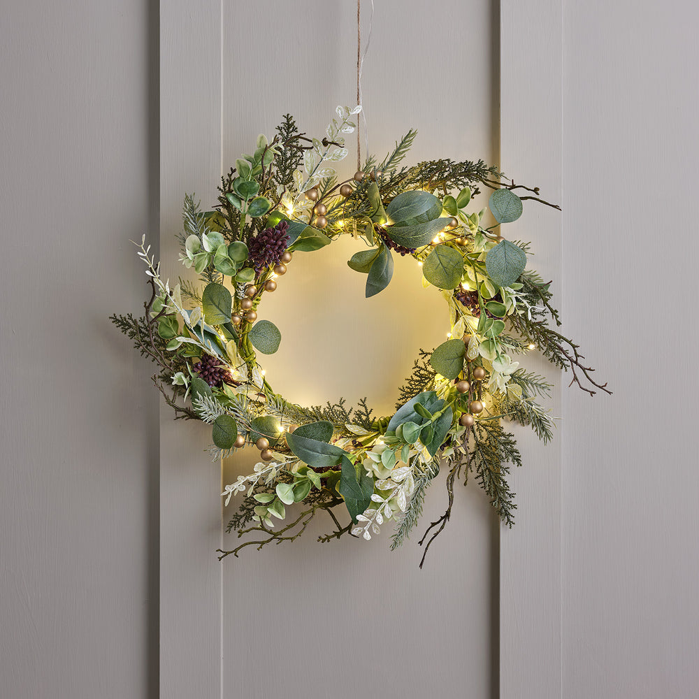 Weihnachtskranz mit grünen Blättern, goldenen Beeren und warmweißer Micro-LED-Lichterkette an grauer Wand