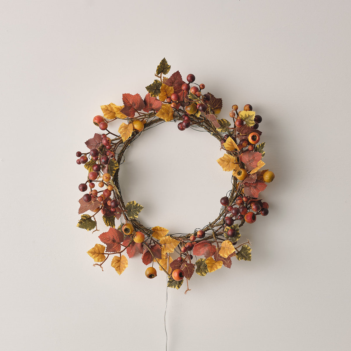 Herbstkranz mit roten und gelben Blättern und Beeren ohne eingeschaltete Lichterkette an weißer Wand