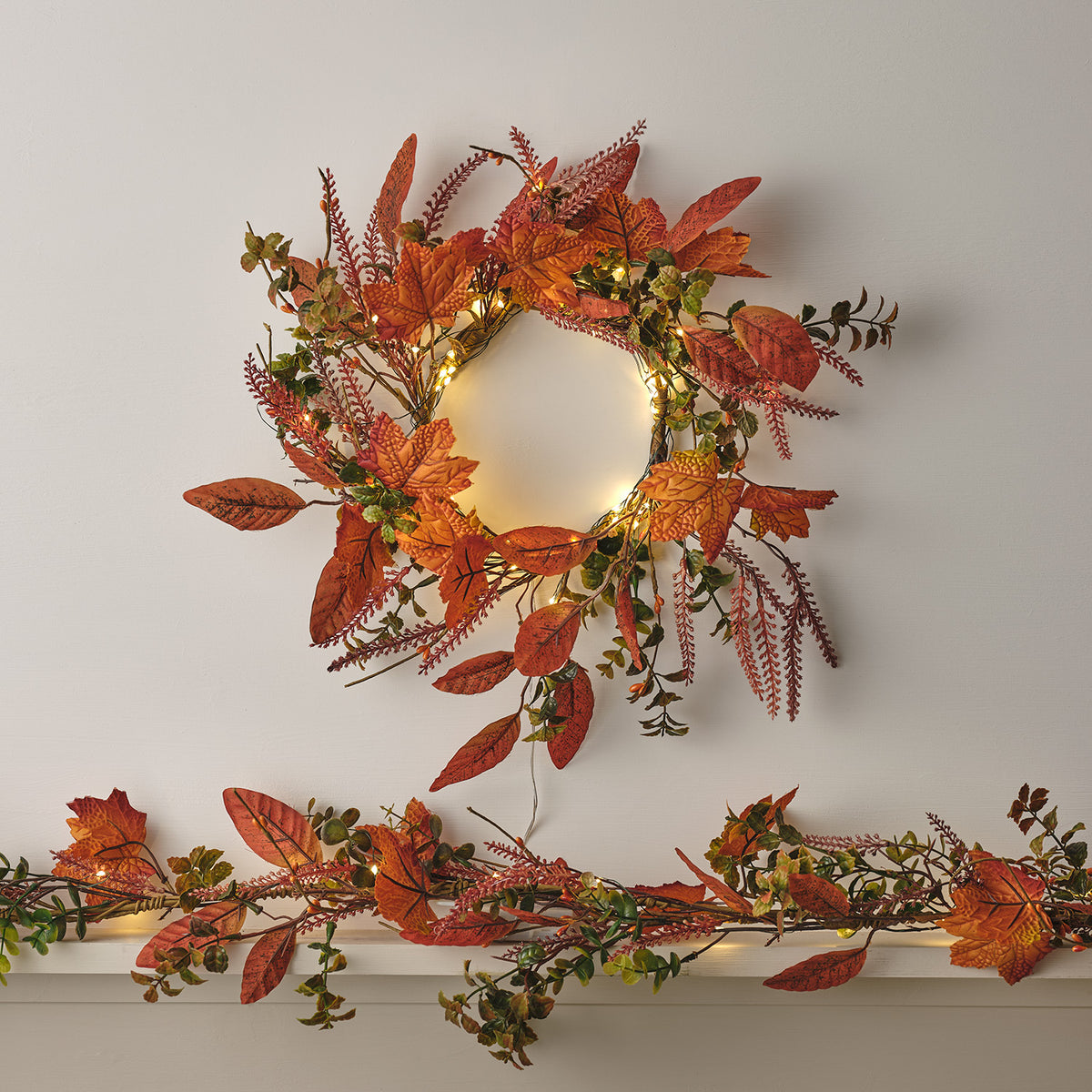Herbstkranz mit orangefarbenen Ahornblättern und kleinen Lichterketten an weißer Wand