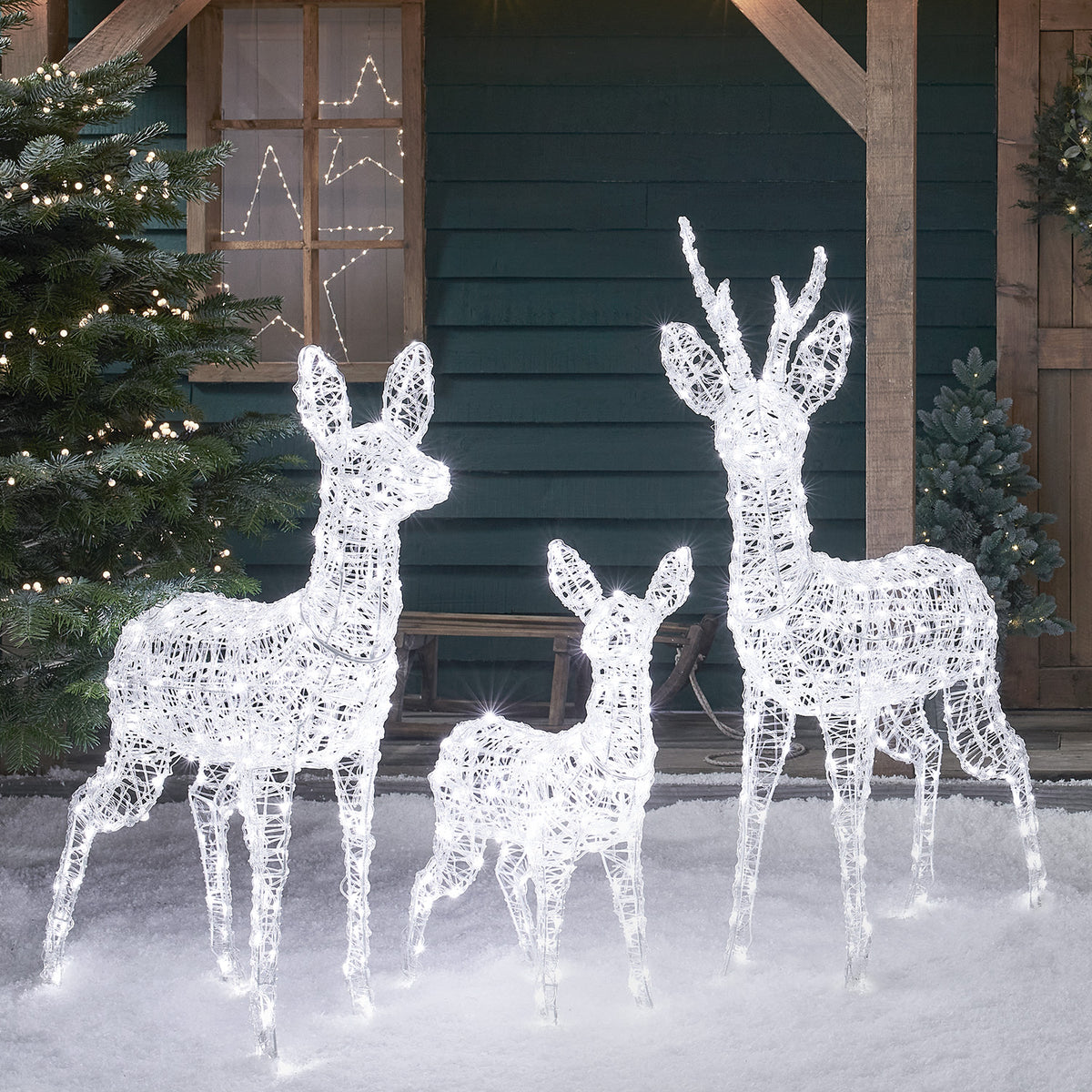 Drei weiße LED-Rentierfiguren aus Acryl auf Schnee vor grünem Holzhaus mit Tür und Fenster