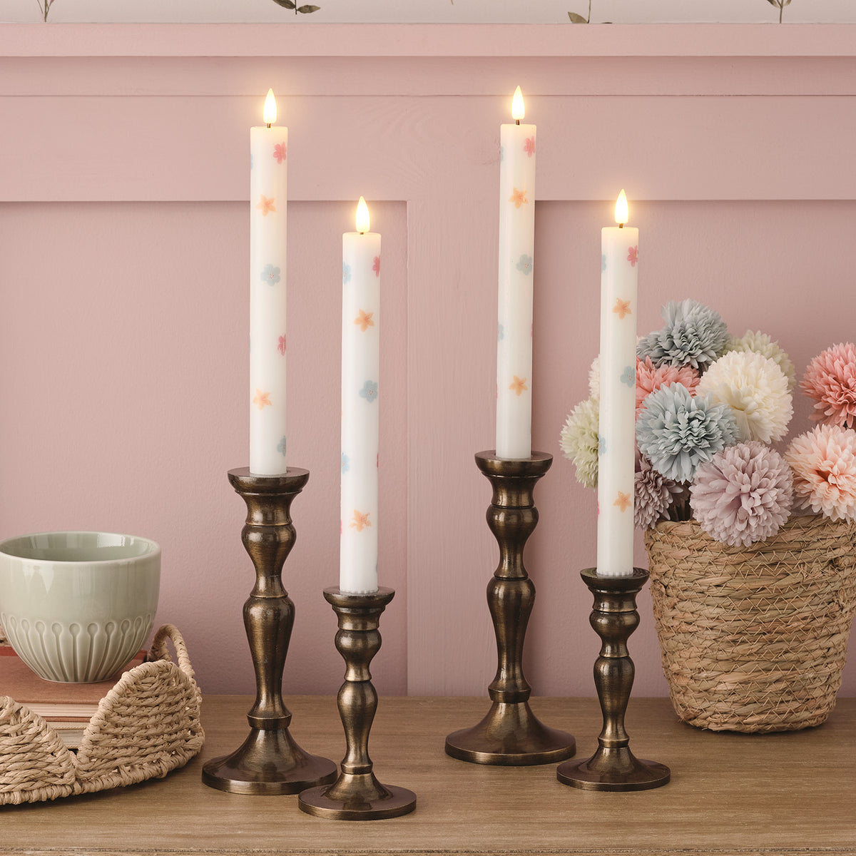 Vier weiße LED Stabkerzen mit pastellfarbenen Blumen in dunklen Kerzenhaltern vor rosa Wand