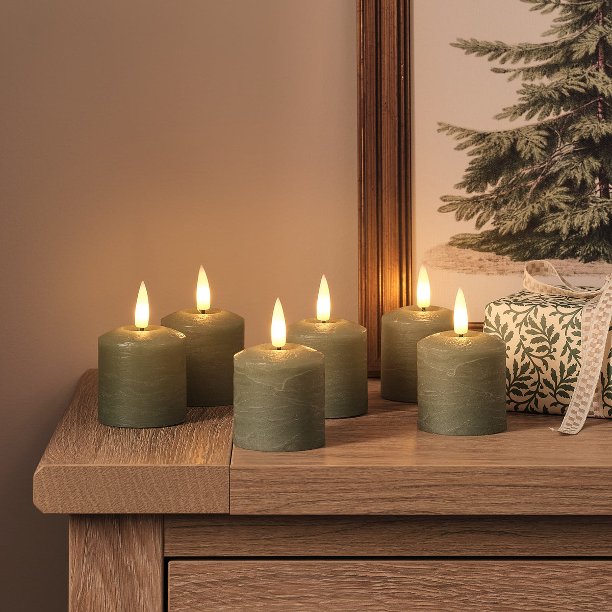 Sechs grüne LED-Votivkerzen mit warmweißem Licht auf Holzkommode vor Weihnachtsbild