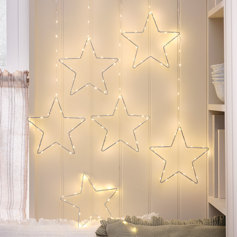 Sechs silberne LED-Sterne mit warmweißem Licht hängen vor einer hellen Wand