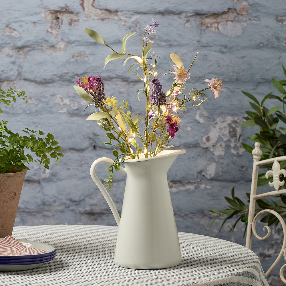 Bunte Frühlingszweige mit warmweißen LED-Lichtern in weißem Krug auf gestreiftem Tischtuch