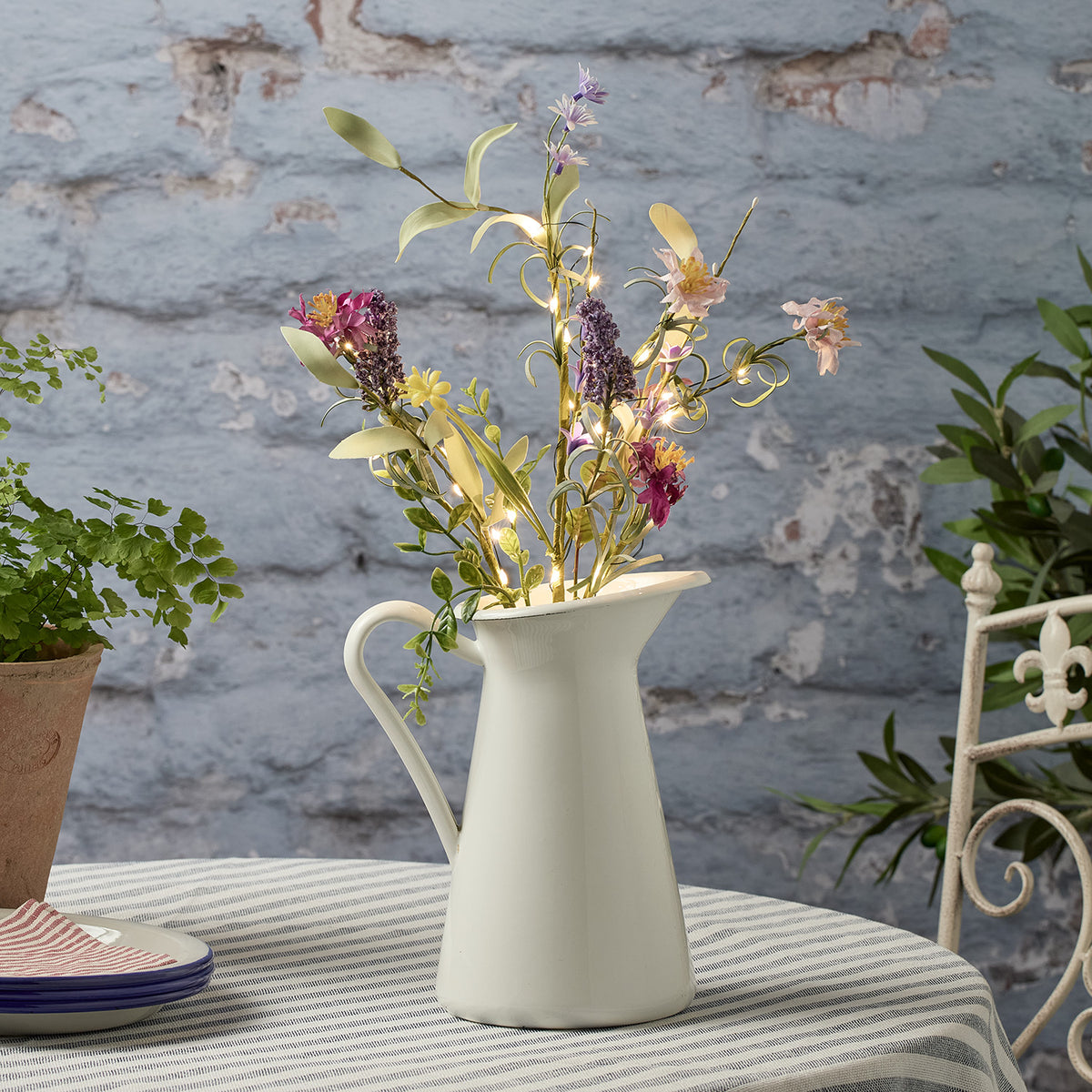 Bunte Frühlingszweige mit warmweißen LED-Lichtern in weißem Krug auf gestreiftem Tischtuch
