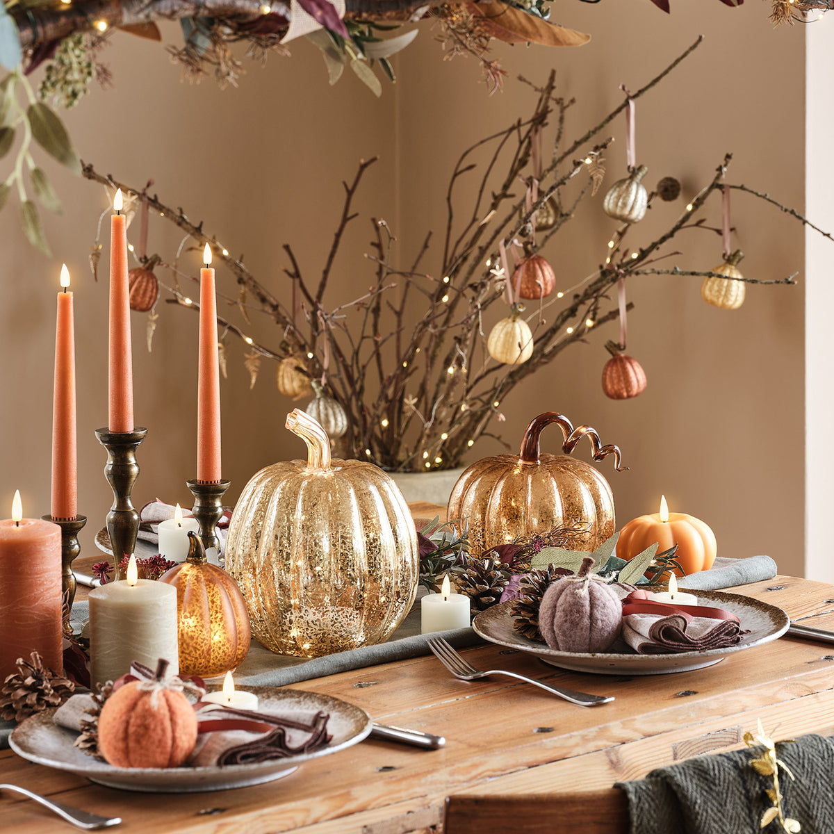 Herbstlich gedeckter Tisch mit Kerzen, Stoffkürbissen und Glas-Kürbissen vor Zweigen mit Lichterkette