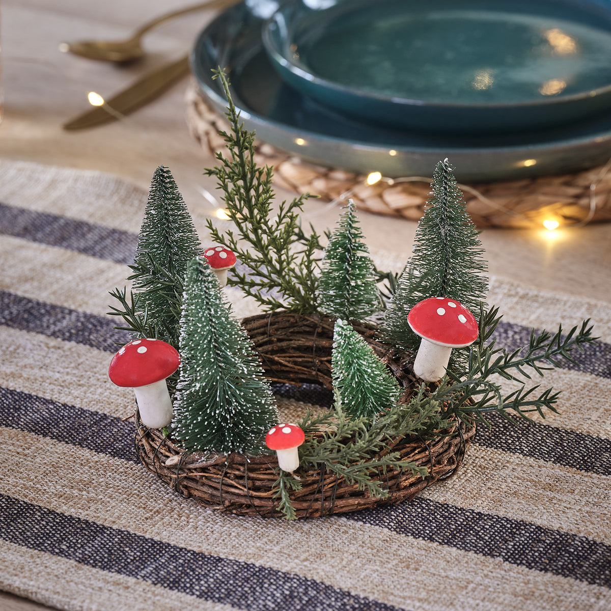 Runder Rattan-Kerzenhalter mit kleinen roten Pilzen und Mini-Tannen auf Tischdecke