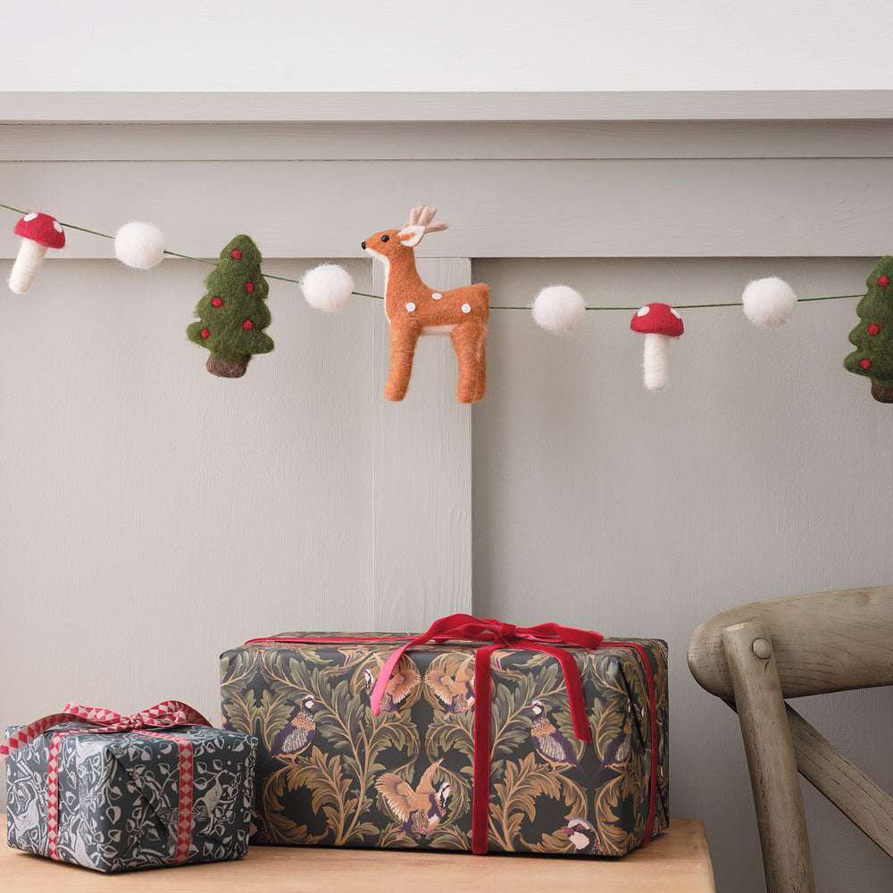 Weihnachtsgirlande aus Filz mit Rentier, Tannenbäumen, Pilzen und weißen Kugeln über einem Tisch mit Geschenkpaketen