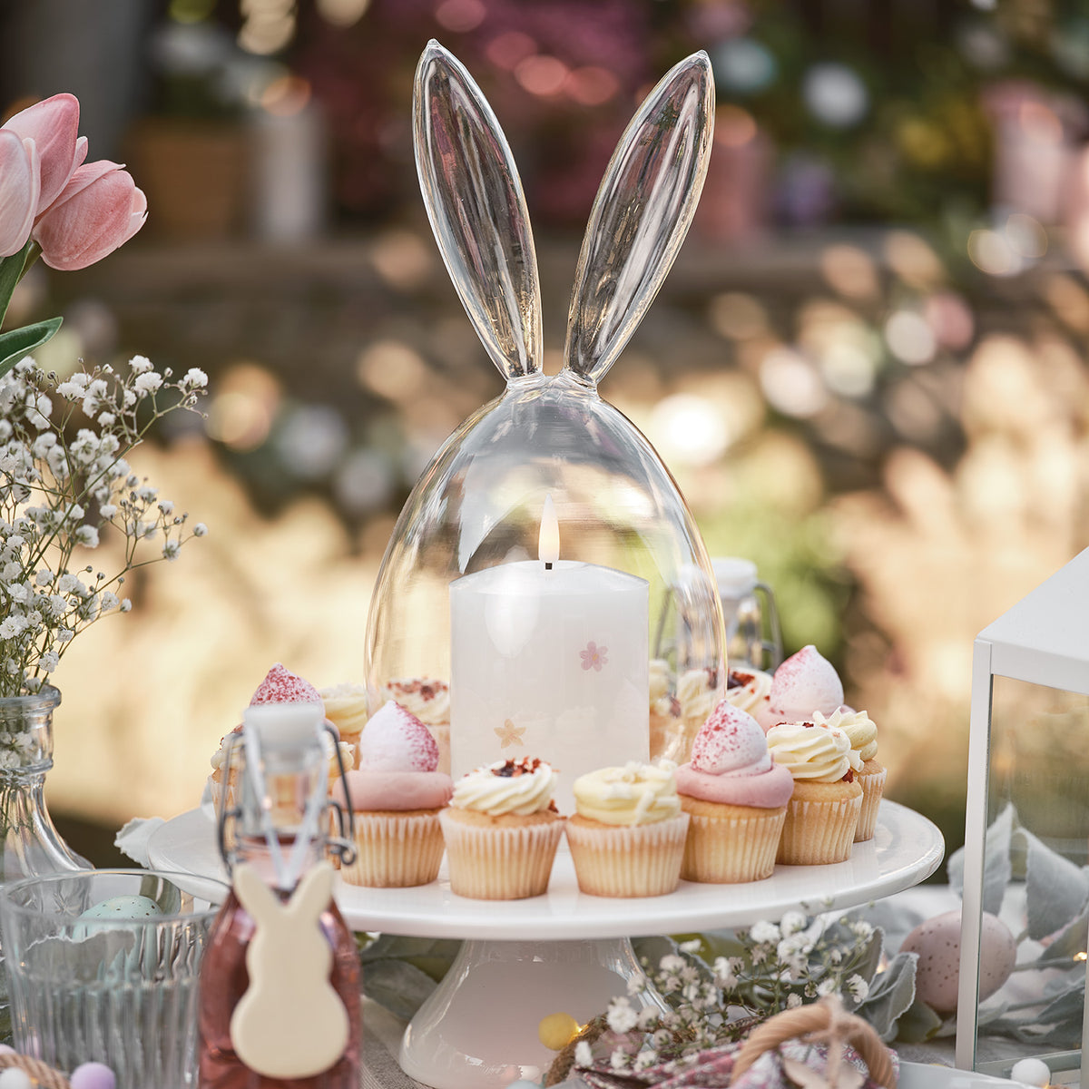 Glaskuppel mit Hasenohren auf weißem Kuchenständer mit brennender Kerze und Cupcakes
