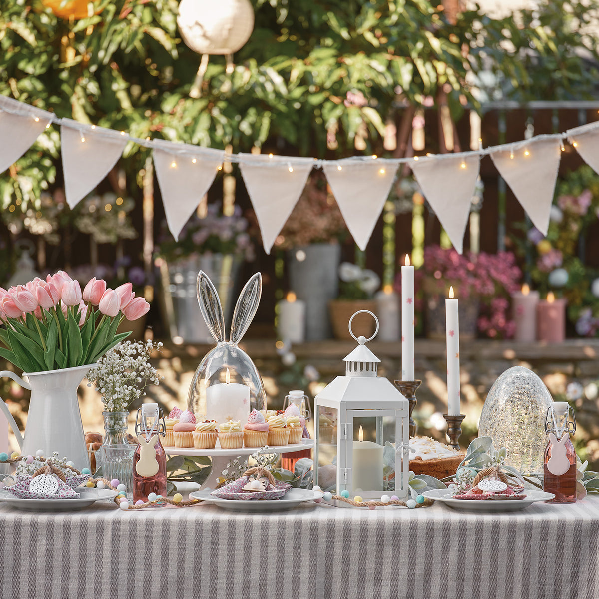 Ostertisch mit Glaskuppel-Hasen, Kerzen, Blumen und Dekoration im Garten