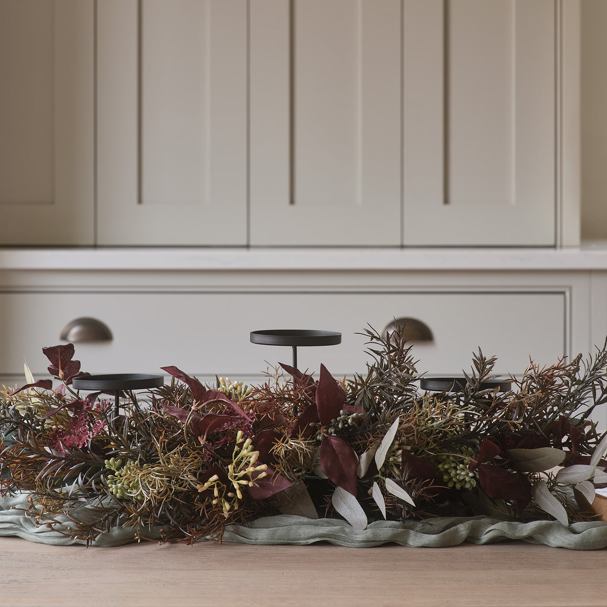 Herbstlicher Kerzenhalter mit drei schwarzen Metallkerzentellern und Kunstlaub vor weißem Schrank auf Holztisch