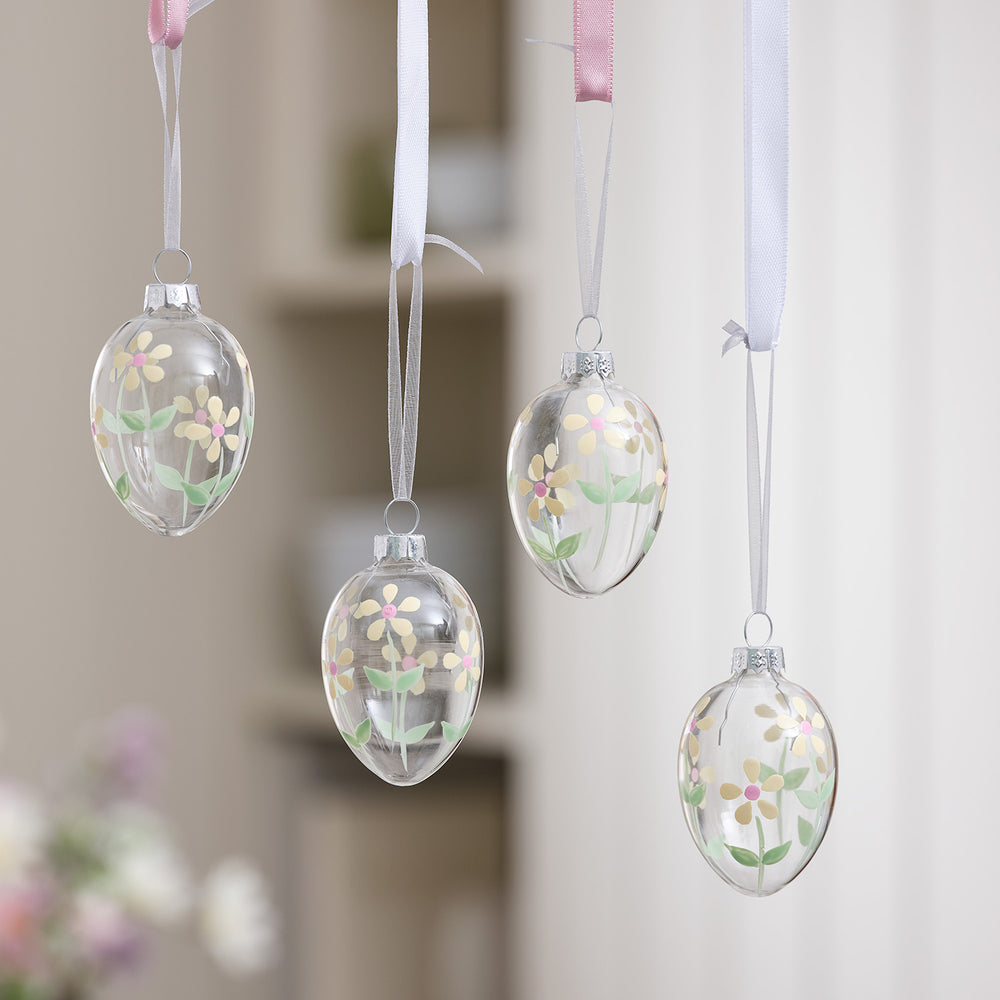 Vier handbemalte Glas-Ostereier mit Blumenmotiv hängen an weißen und rosa Bändern