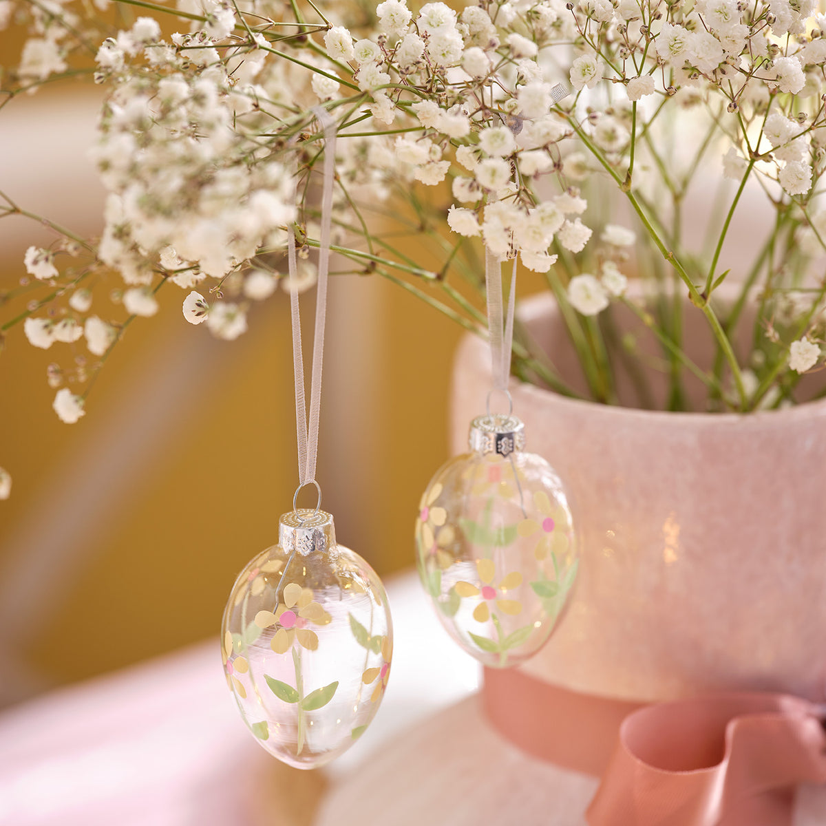 Zwei Glas-Ostereier mit Blumenmotiv hängen an weißen Bändern vor einem rosa Blumentopf