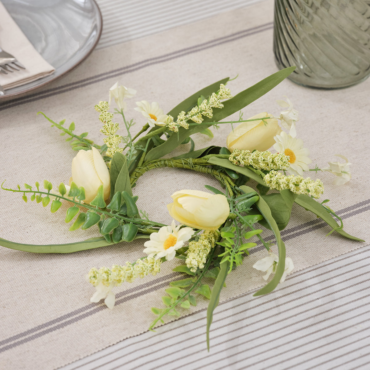 Kerzenring mit buttergelben Tulpen, Gänseblümchen und grünen Gräsern auf Tischdecke