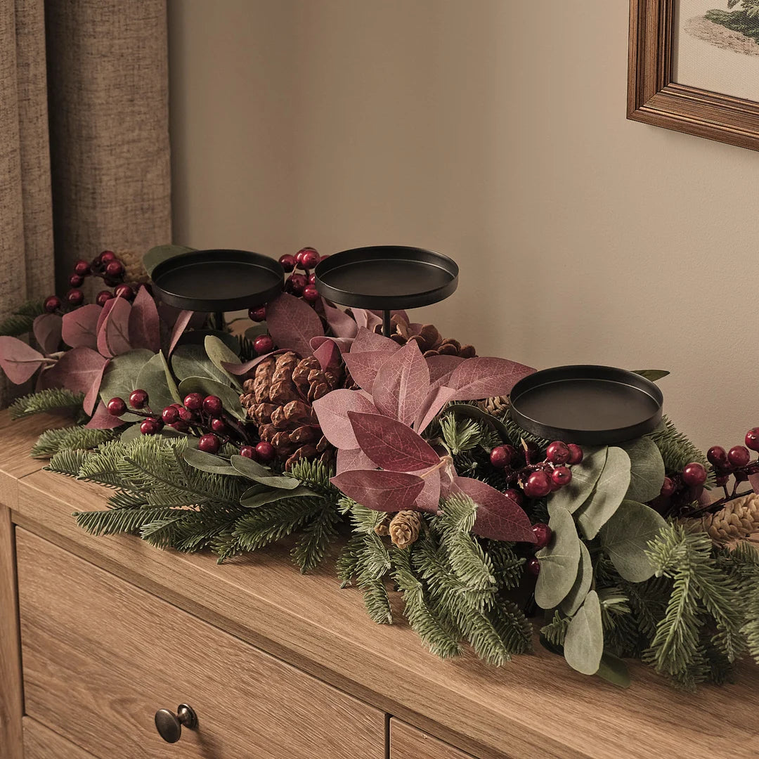 Elegantes Adventsgesteck mit drei schwarzen Kerzenhaltern auf natürlichem Holztisch, dekoriert mit burgundy-farbenen Blättern, grünem Fichtengrün, roten Beeren, braunen Zapfen und cremefarbenen Akzenten – festliche Tischdekoration für klassische Weihnachtsstimmung und edle Wohnzimmer-Arrangements.