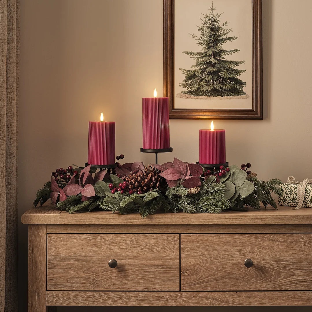 Detailaufnahme eines hochwertigen Adventsgestecks mit drei burgundy-roten LED-Taperkerzen mit warmweißem Flammeneffekt in schwarzen Kerzenhaltern, arrangiert auf Holzkommode mit natürlichem Fichtengrün, burgundy Akzent-Blättern, roten Beeren, braunen Pinienzapfen und cremefarbenen Dekorfiguren – luxuriöses Adventsarrangement für stilvolle Kamindekoration und gehobene Festtischlooks.