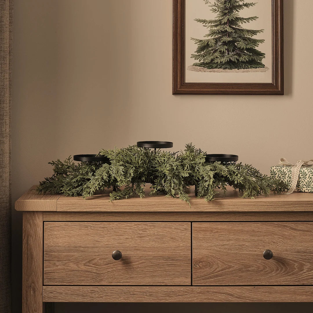 Stilvolles Adventsgesteck auf Holzkommode mit schwarzen Kerzenhaltern, dichtem grünem Fichtengrün-Arrangement, cremefarbenen Dekoelementen und gerahmtem Tannenbaum-Kunstdruck im Hintergrund – klassische edle Weihnachtsdekoration für gehobene Raumszenen und festliche Kaminsims-Displays.