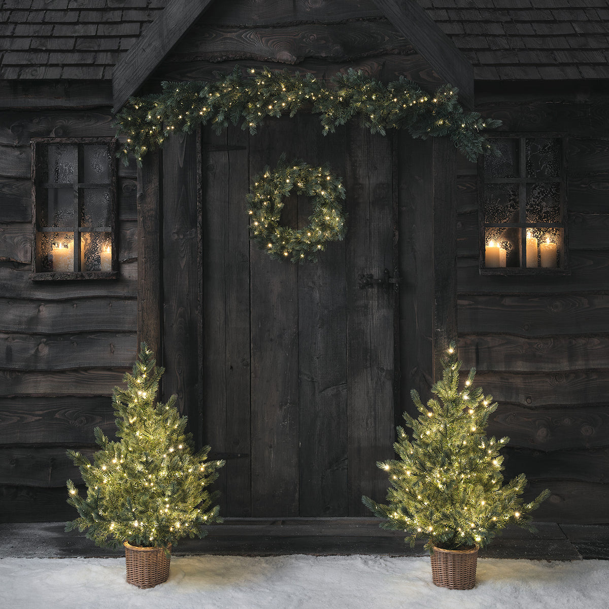 Weihnachtskranz, Girlande und zwei beleuchtete Weihnachtsbäume vor dunkler Holztür