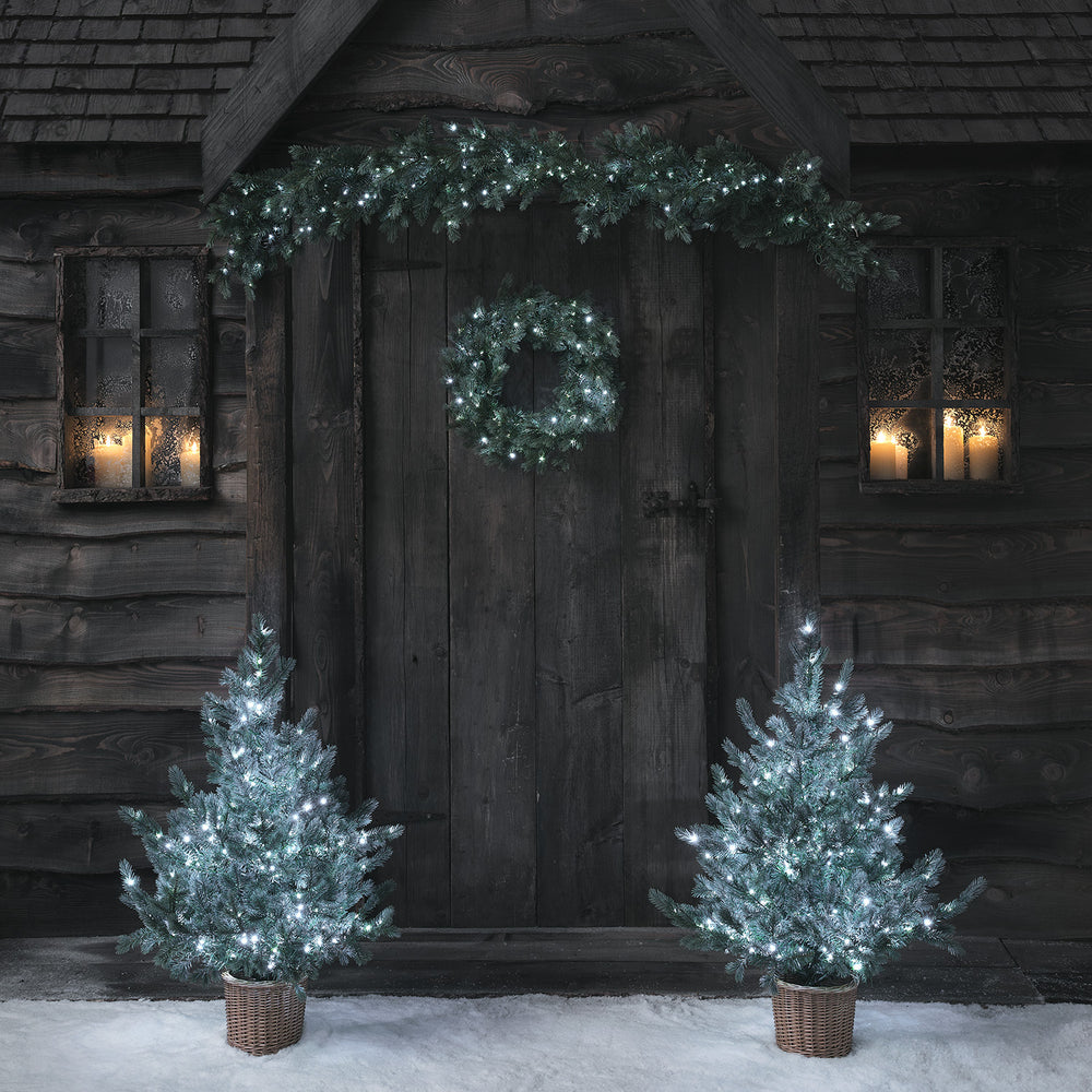 Weihnachtskranz, Girlande und zwei Weihnachtsbäume mit kaltweiß leuchtenden LEDs vor Holztür