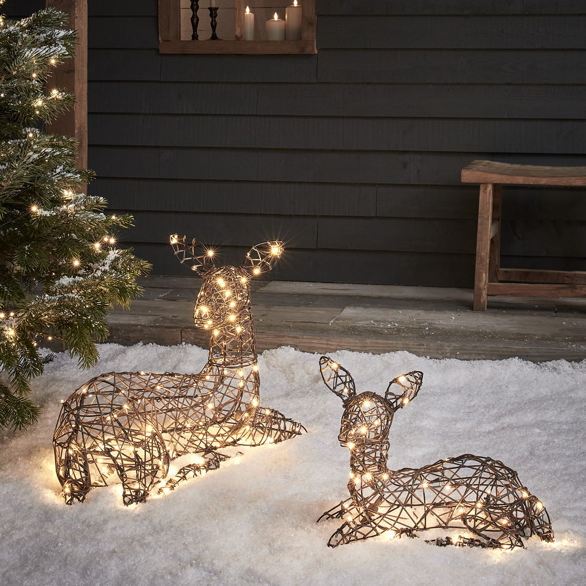 Liegende LED-Rentiere Mutter und Kind aus braunem Rattan mit warmweißen Lichtern im Schnee vor dunkler Wand
