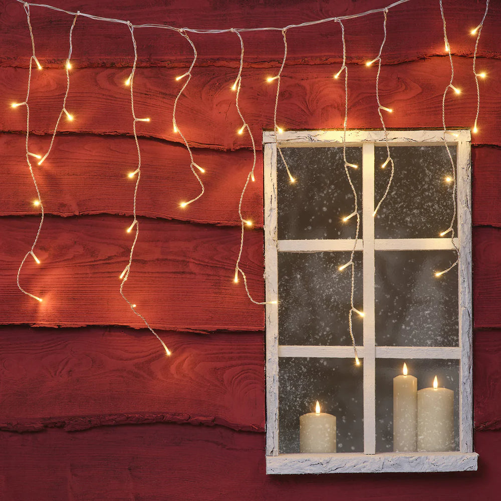 Warmweiße Eiszapfen-Lichterkette mit 100 LEDs hängt vor roter Holzfassade neben Fenster mit brennenden Kerzen