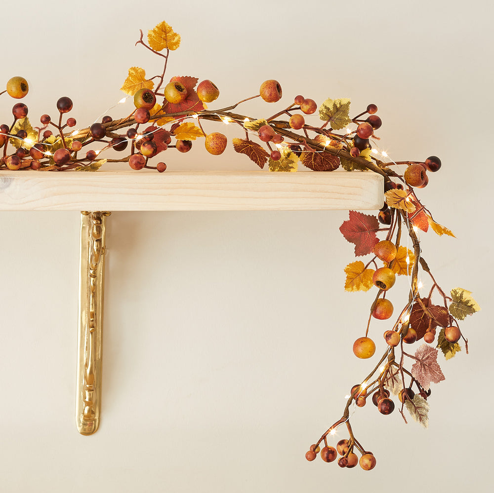Herbstliche Girlande mit Beeren, bunten Blättern und kleinen Lichterketten auf einem Holzregal