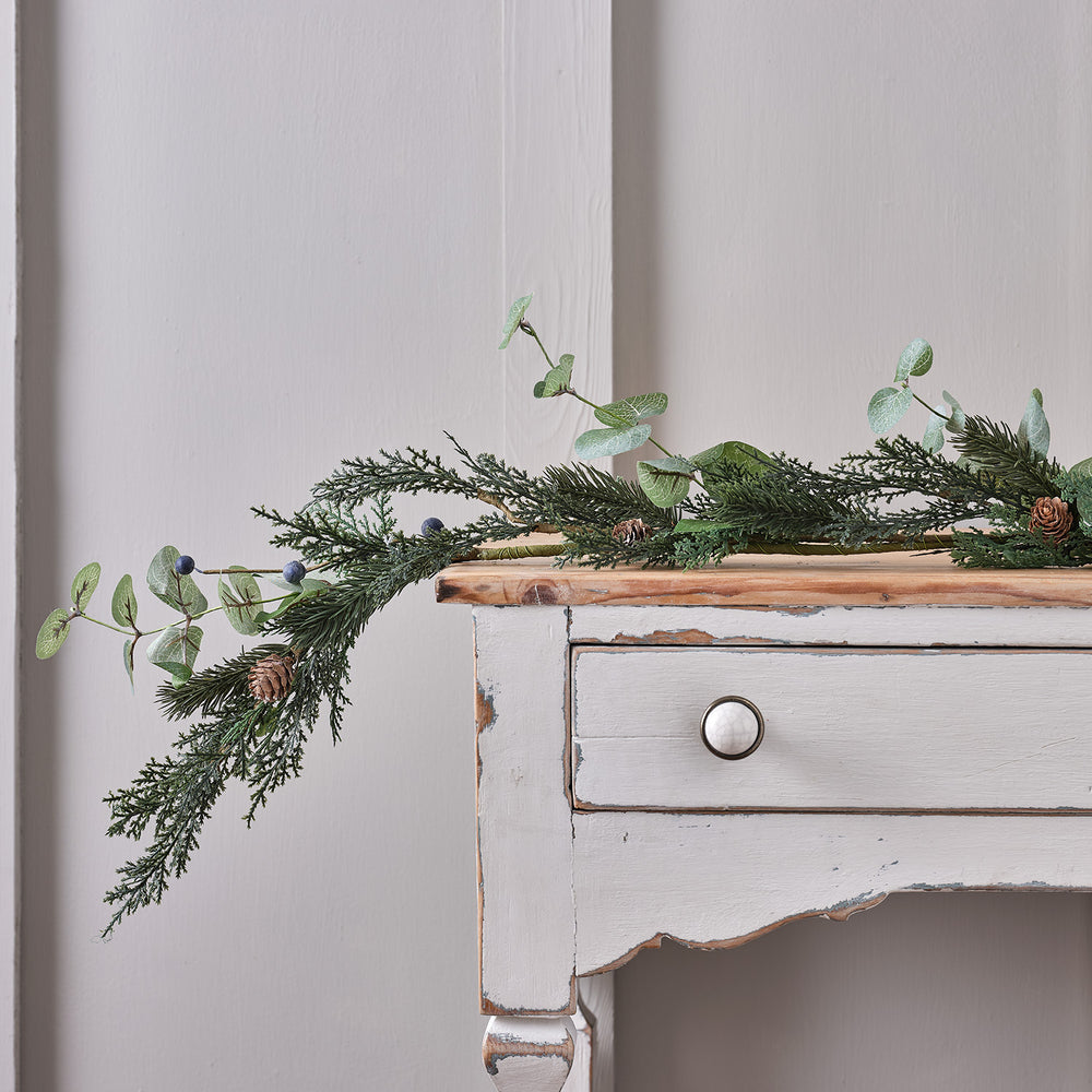 Weihnachtsgirlande mit grünen Blättern, Tannenzapfen und blauen Beeren auf weißem Holztisch
