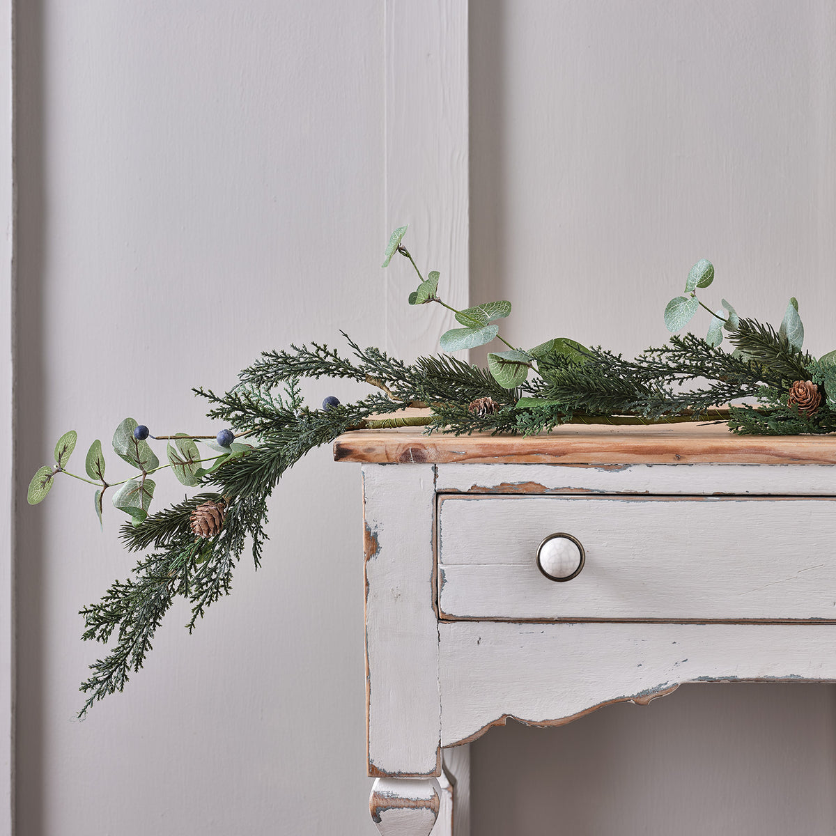 Weihnachtsgirlande mit grünen Blättern, Tannenzapfen und blauen Beeren auf weißem Holztisch