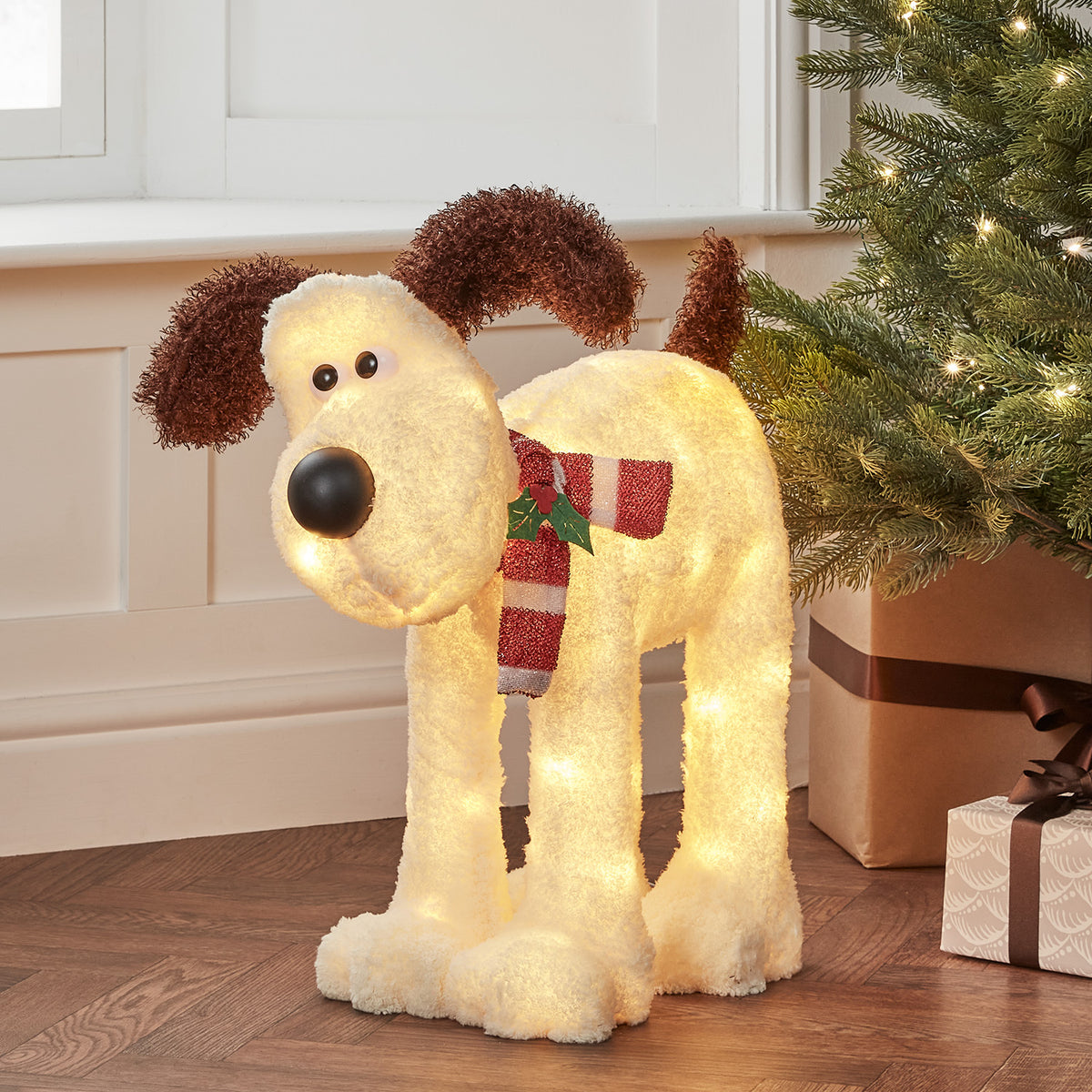 Gromit LED-Figur mit rot-weißem Schal neben Weihnachtsbaum und Geschenken im Innenraum