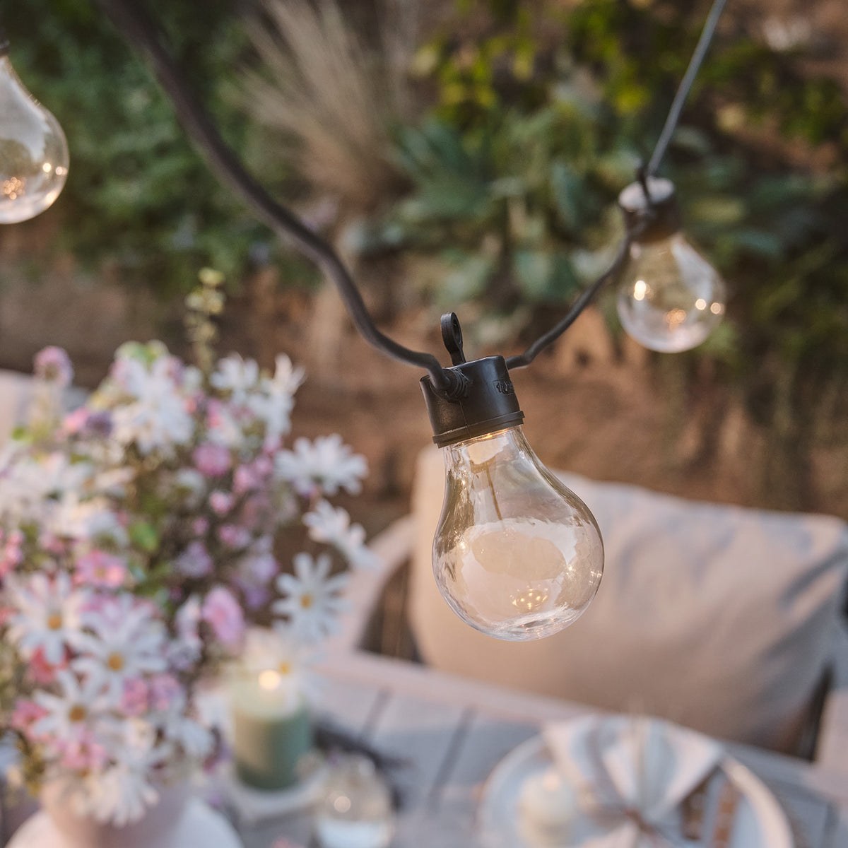 Detailansicht einer warmweißen Glühbirne der Lichterkette mit Blumen im Hintergrund