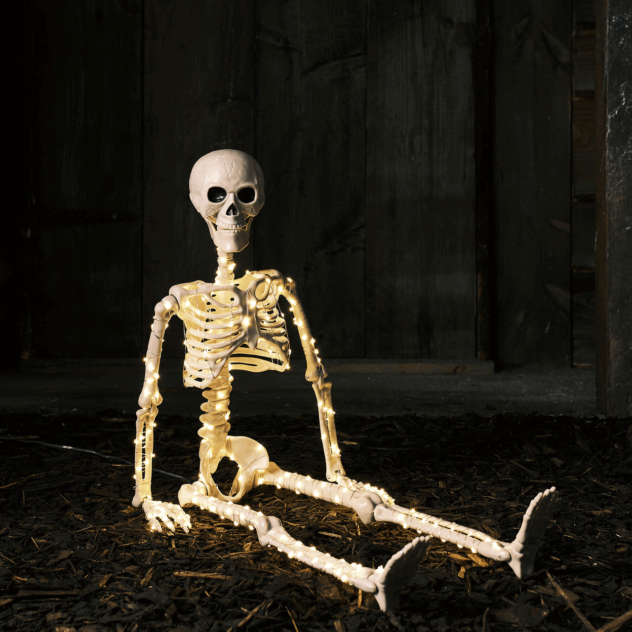Skelett mit Micro LEDs sitzt auf dunklem Boden vor dunkler Holzwand, Beine ausgestreckt