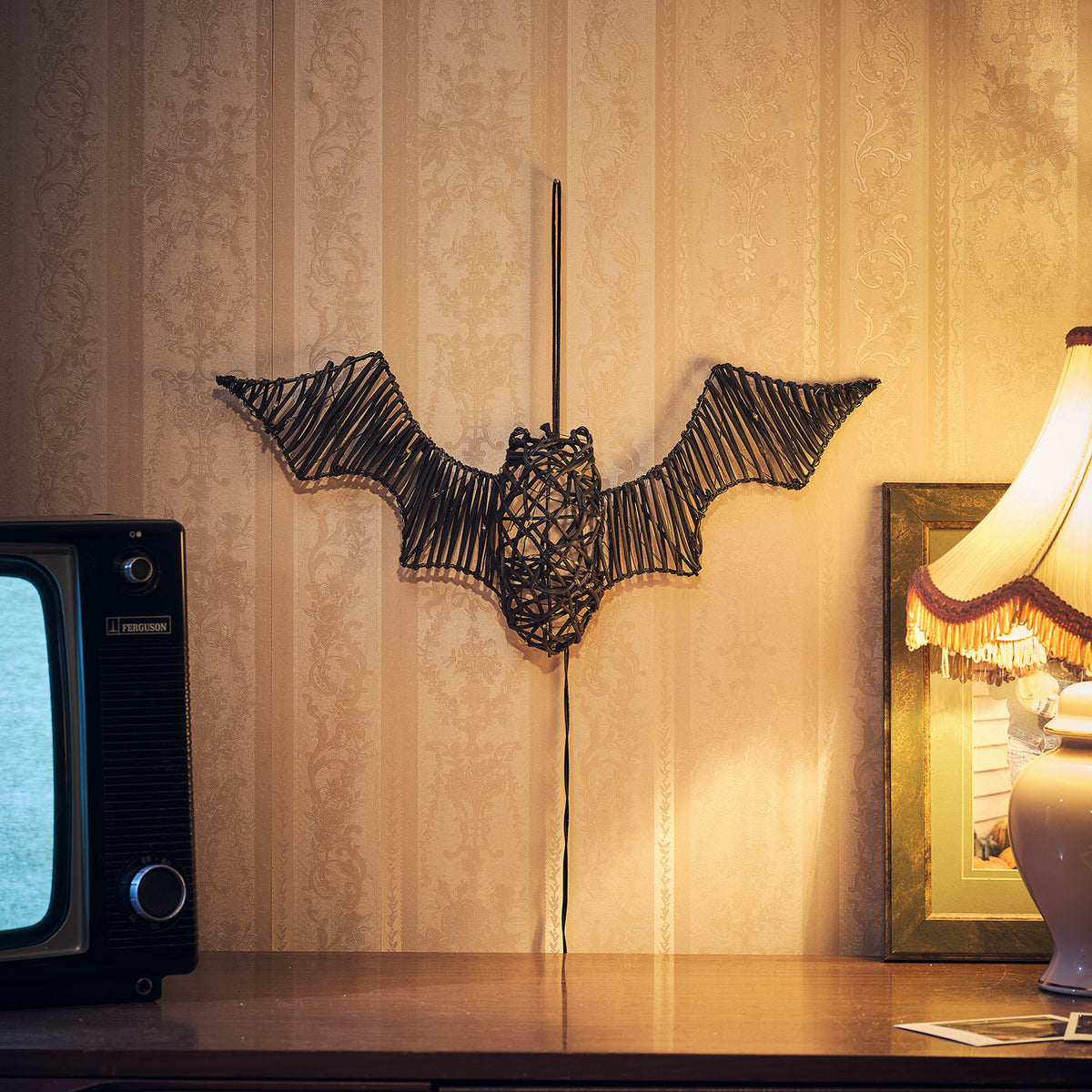 Unbeleuchtete Rattan-Fledermaus an Wand über Tisch mit Lampe und Bilderrahmen