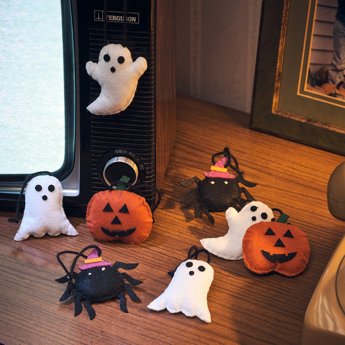 Halloween-Filzanhänger Kürbis, Geist und Spinne liegen auf Holzkommode neben altem Fernseher