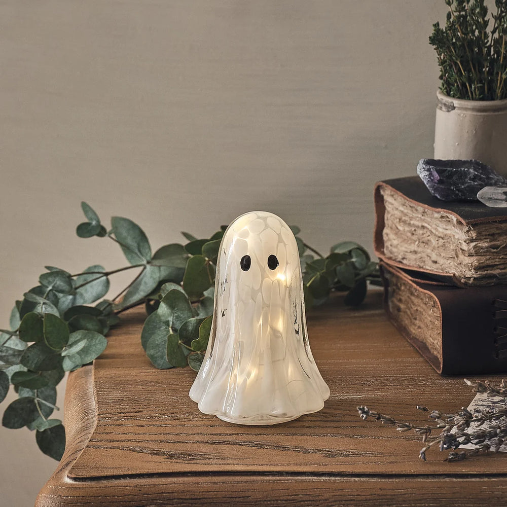 Kleine LED-Halloween-Geist-Deko aus Konfettiglas auf Holztisch mit Pflanzen im Hintergrund
