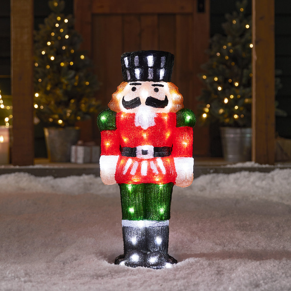 Beleuchtete Acryl-Nussknacker-Figur in Rot, Grün und Schwarz steht im Schnee vor einer Haustür mit Weihnachtsbäumen