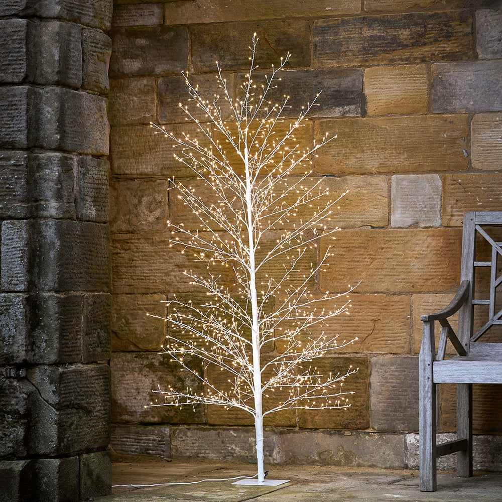Weißer Draht-Baum mit warmweißen LED-Lichtern vor einer Steinmauer und neben einer Holzbank