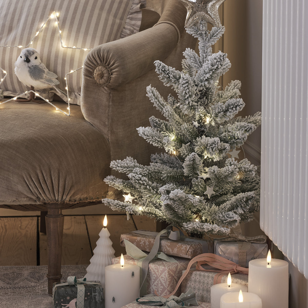 Verschneiter Mini-Weihnachtsbaum mit Lichterkette neben braunem Sessel und Kerzen