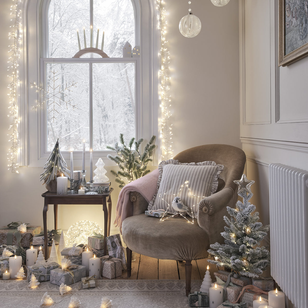 Weihnachtsdeko mit verschneitem Baum, Geschenken, Kerzen und Lichterketten vor Fenster