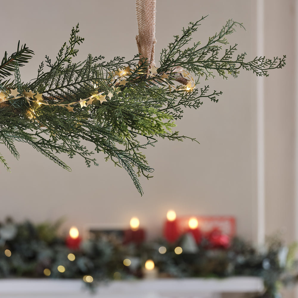Nahaufnahme einer künstlichen Zypressen-Weihnachtsgirlande mit Sternen-Lichterkette vor Kerzenlicht
